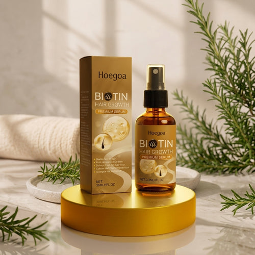 Hoegoa Biotin Hair Growth Premium Serum bottle and matching box displayed on a gold pedestal with spa-style props including a towel, marble surface, and greenery.